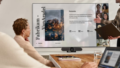 Persone in una sala riunioni che guardano una presentazione su un grande televisore durante una videochiamata di Teams.