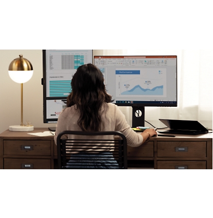 Osoba, která sedí v domácí kanceláři a dívá se na data na dvou stolních monitorech.