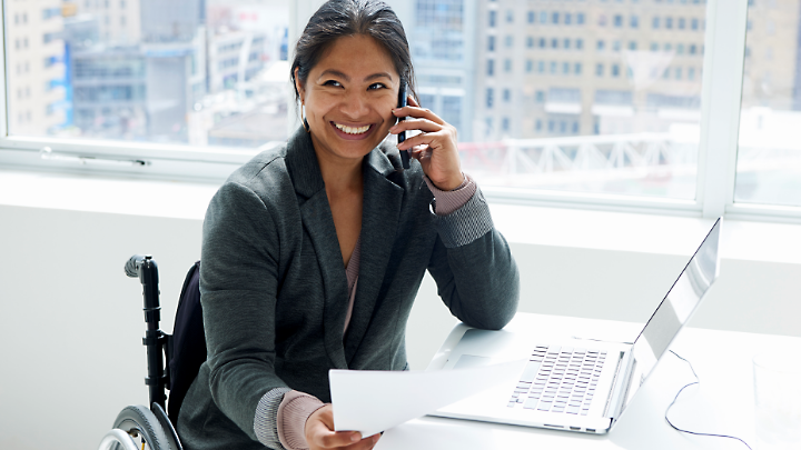 A customer support representative in a wheelchair smiling, sitting at a desk with a laptop and holding a mobile phone to their ear.