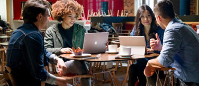 ארבעה אנשים יושבים בשולחן קפה, שניים מהם מחשבים נישאים, נראים מעורבים בדיון. 