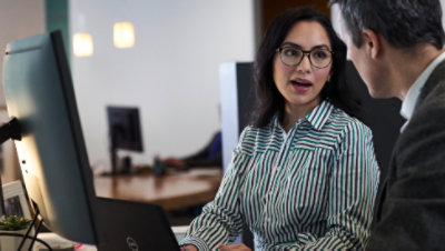 Two people talking while seated at a desk with a display monitor and a laptop