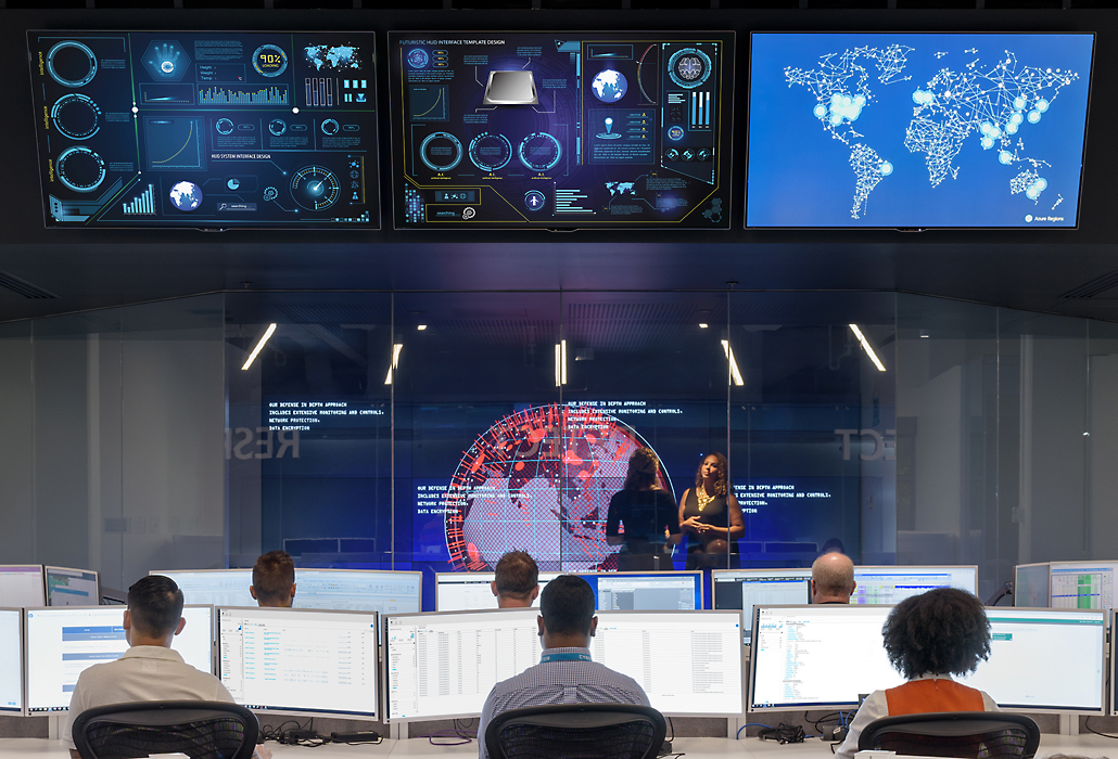 Individuals in a data center monitoring data flow in front of larger hanging monitors