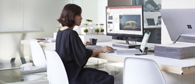 Una persona lavora al computer su una scrivania in un ufficio con documenti, un portatile e una pianta vicino.