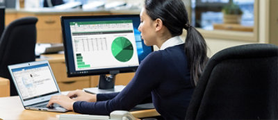 Kobieta siedzi przy biurku w biurze, pracując na laptopie, podczas gdy monitor komputera przed nią wyświetla arkusz kalkulacyjny z zielonym wykresem kołowym.