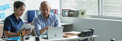 Due persone lavorano alla scrivania con un portatile e documenti. Sono concentrati sugli schermi e sui fogli.