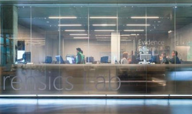 View through a glass wall with the words Forensics Lab shows a room where people are working on computers