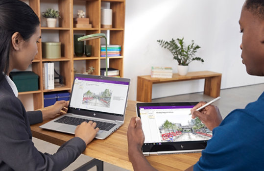 Fotografía de dos personas sentadas ante un escritorio. Una está trabajando con un portátil. Una está trabajando en una tableta con un lápiz.