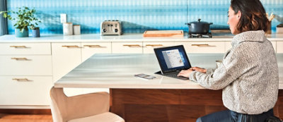 Persona seduta al tavolo della cucina che usa un portatile. Sullo sfondo un tostapane, una pentola sul piano cottura e altri articoli da cucina.