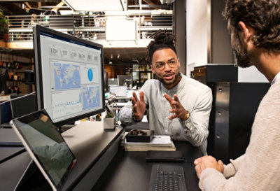 Two people having a conversation at a desk with a desktop monitor displaying maps and data