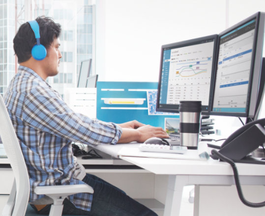 A person working in an office. They are wearing headphones and are seated at a desk with two display monitors.