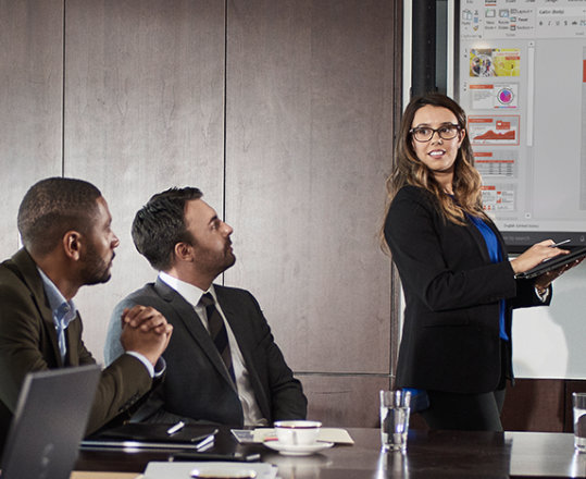 Photograph of a group of people seated at table in a conference room. There is a person presenting in front of a large display monitor.