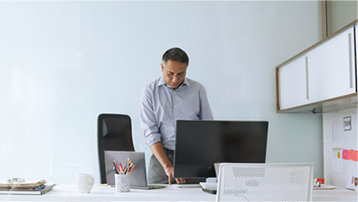 A person standing at a desk in an office working. There is a laptop and a display monitor on the desk.