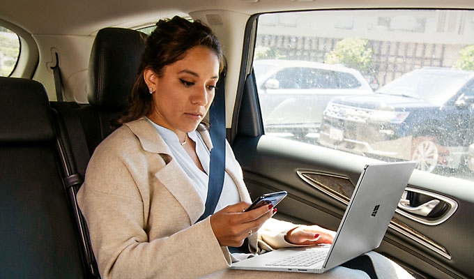 A person sitting in the back seat of a car using a laptop and looking at a mobile phone