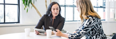 Dve žene sede za stolom u kancelariji i razgovaraju, a svaka od njih drži tablet i pametni telefon