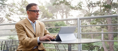 Un uomo che indossa una giacca beige lavora al tablet su un tavolino all'aperto sotto un patio, circondato da alberi.