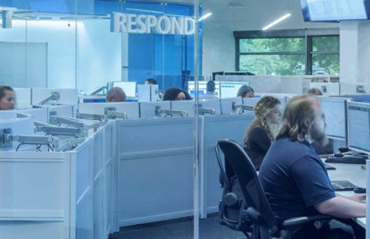 View from a hallway looking through security professionals working on computers in a room with glass walls.