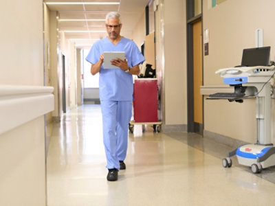 A person wearing blue scrubs walks in a hospital hallway near a machine with a blue and white device.
