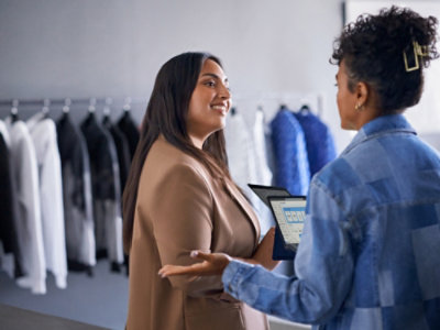 A woman in a brown jacket holding a tablet while talking to another person.