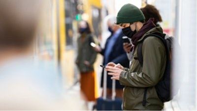A person wearing PPE looking at a mobile phone while waiting for public transportation