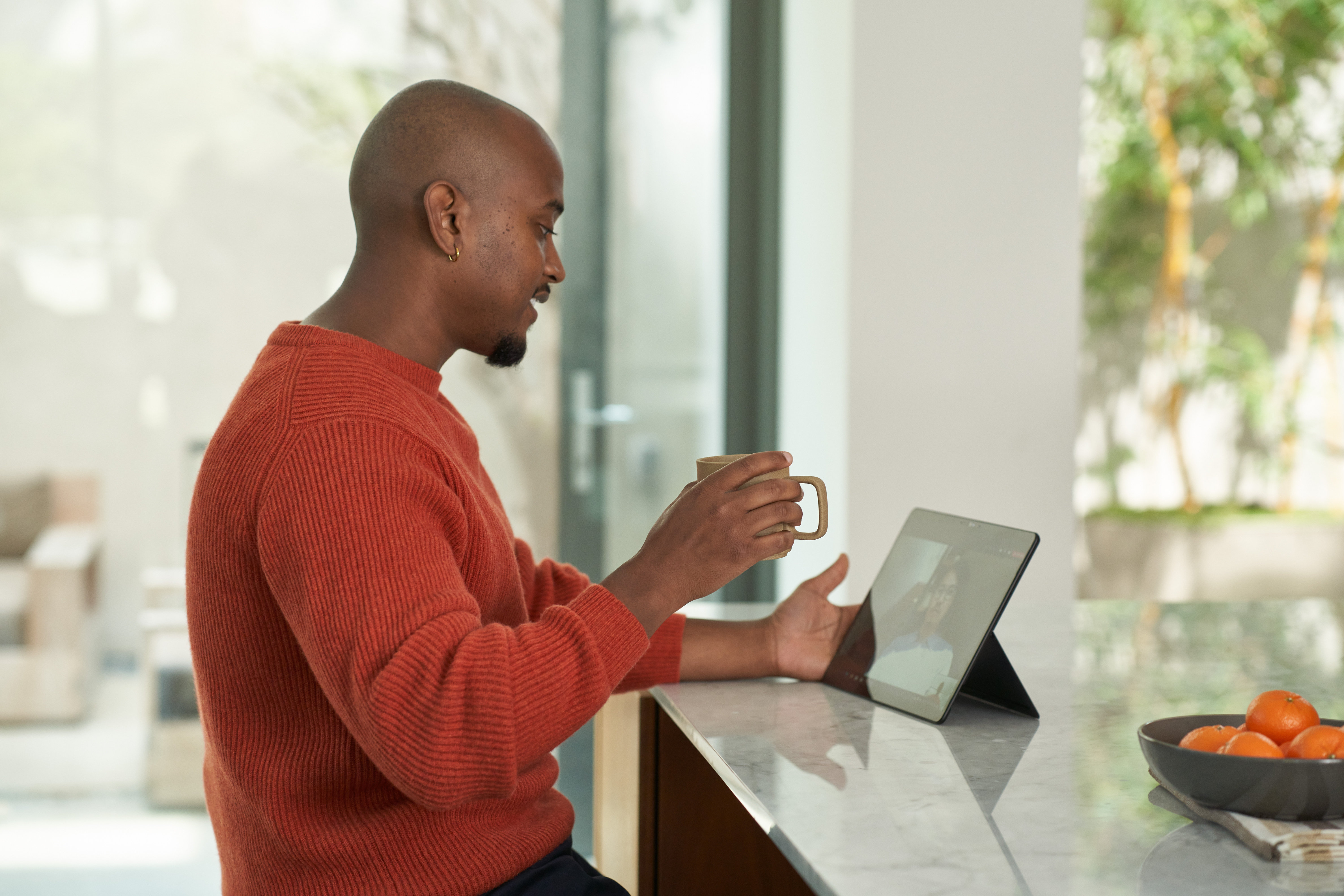 A smiling man in a red sweater sits in an open room holding a coffee mug and watching a tablet screen.