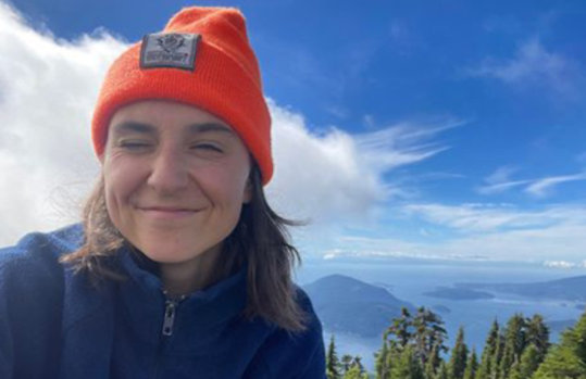 Kiah McGeer wearing an orange beanie and a blue jacket stands in front of a landscape featuring green mountains, a lake, and a clear sky