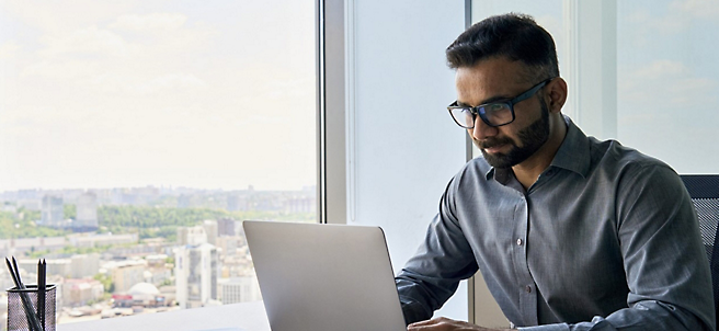 Um homem de óculos trabalhando em um laptop em um escritório com vista para a cidade através das grandes janelas.