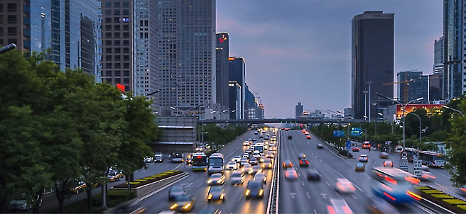 Traffic flows on a multilane city road at dusk with modern skyscrapers lining the sides under a cloudy sky.