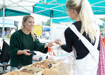 Žena za pijačnom tezgom ispod plavo-bele nadstrešnice pruža kupcu pecivo u beloj kesi.