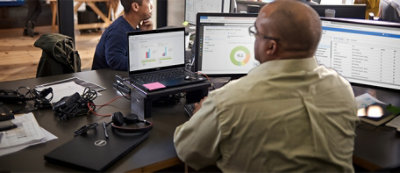 Osoba pracuje przy biurku z wieloma ekranami komputerowymi wyświetlającymi dane i wykresy, laptopem i sprzętem biurowym w przestrzeni biurowej na planie otwartym.