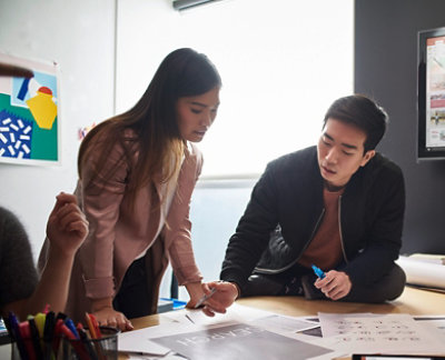 Dve osobe u opremljenoj kancelariji stoje i razgovaraju o štampanim dokumentima na stolu, 