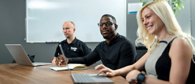 Tri osobe sede za konferencijskim stolom i svaka od njih koristi laptop; jedna osoba u crnoj košulji drži olovku i gleda ka kameri