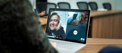 Person participating in a video call shown on a laptop screen, with a woman smiling in the main window and another participant 