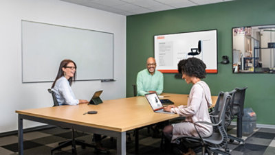 Three people are sitting at a conference table with laptops, engaging in a meeting. A presentation slide is displayed on a wall-mounted