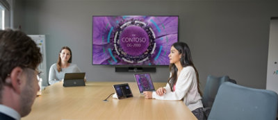 A person sitting at a table with a computer screen and one person in background