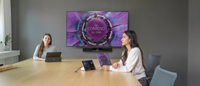 A woman sitting at a table with laptops and a purple circle with white text CONTOSO.