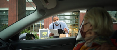 Radnik sa maskom drži tablet i predaje smeđu papirnu kesu osobi u automobilu na lokaciji za preuzimanje ispred zgrade.