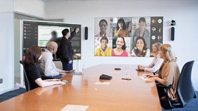 A man stands next to a screen in a conference room, pointing as he presents to both in-person attendees and participants joining through a video call displayed on a large screen.