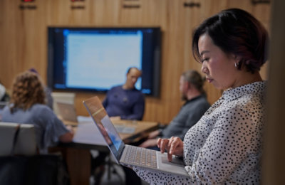 A person standing on the outskirts of a meeting using a laptop 