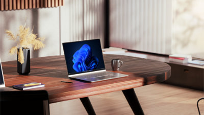 An open laptop displaying a Windows bloom background sits on a wooden desk in a well-lit office, alongside an espresso cup, vase of flowers, a notebook and pencils