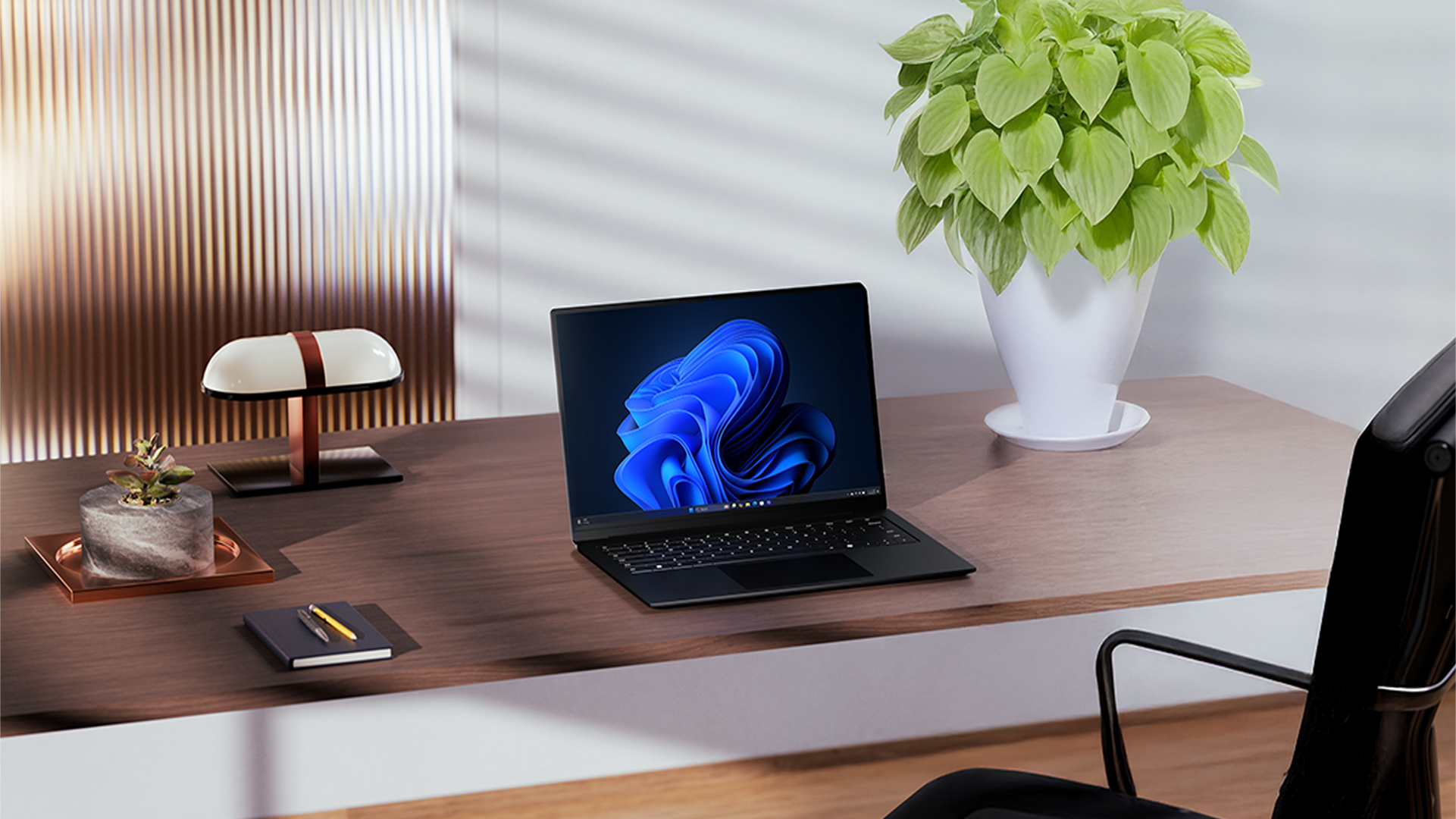 An open laptop displaying a Windows bloom background sits on a wooden desk alongside a lamp, notebook, and plants