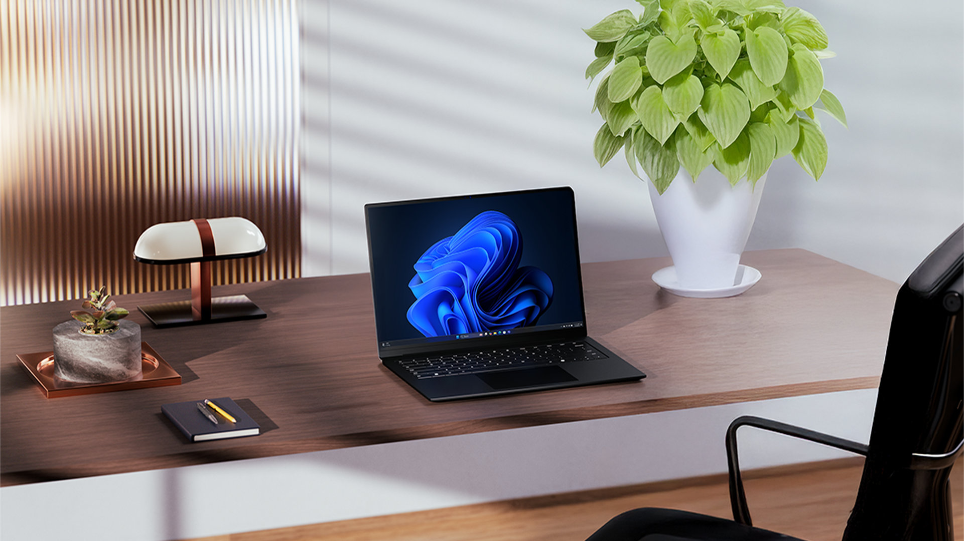 An open laptop displaying a Windows bloom background sits on a wooden desk alongside a lamp, notebook, and plants