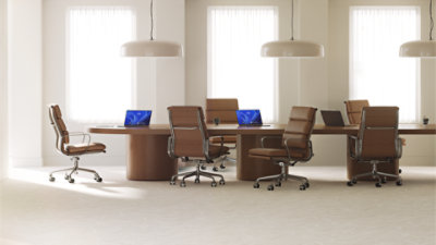 Three open laptops, with two displaying Windows blue bloom backgrounds, sit on a long wood conference room table surrounded by six brown leather office chairs 