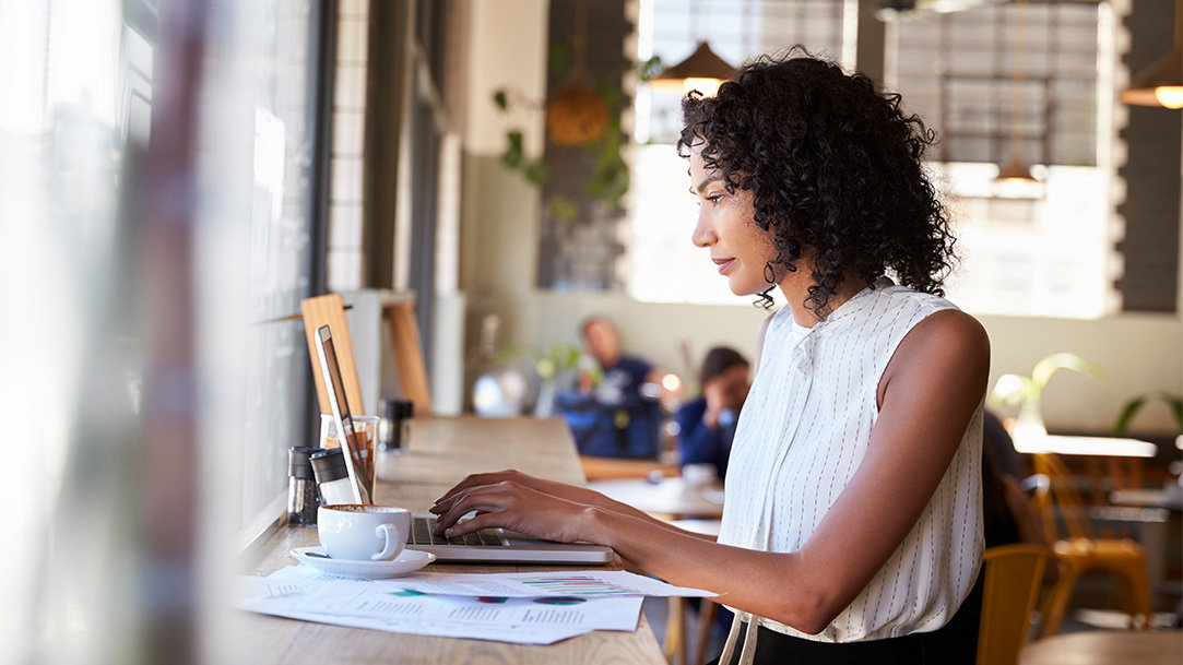 A remote worker sitting at a window bar in a coffee shop working on an open Windows 11 Pro device with notes, a coffee cup, and other tables with customers in the background
