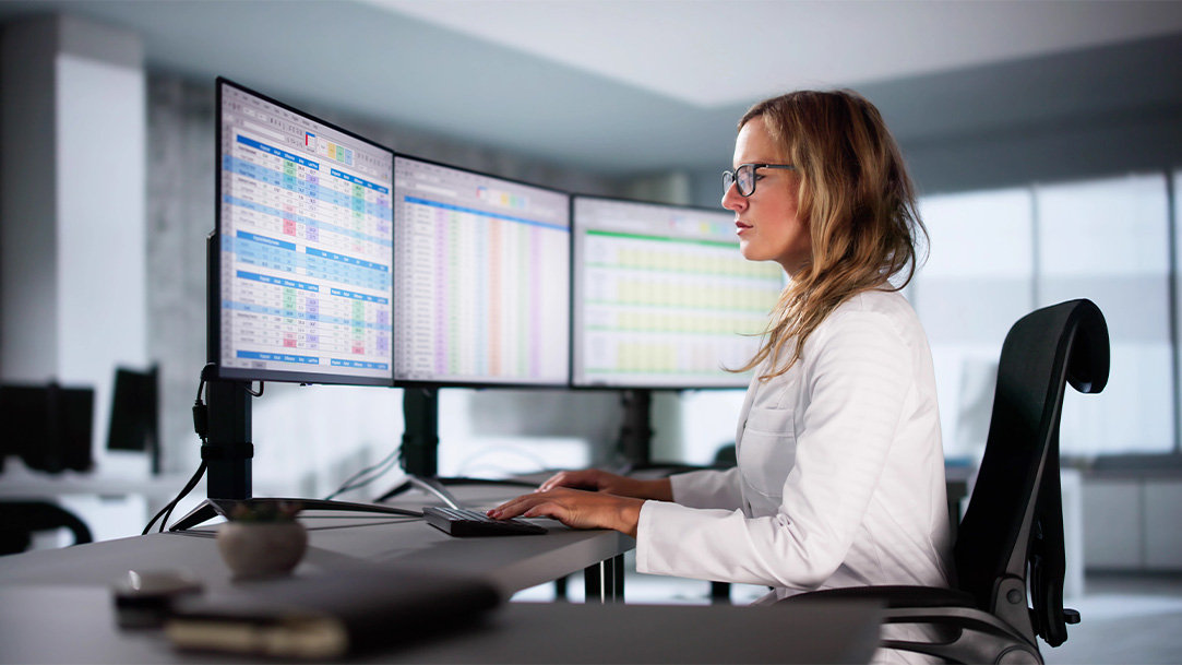 Healthcare worker in a white lab coat sitting at a modern office workstation with three large PC monitors displaying patient data, a keyboard, notebook, and coffee mug