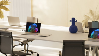 An open laptop displaying a Windows bloom background sits alongside a mouse and notebook on a white desk with a blue chair
