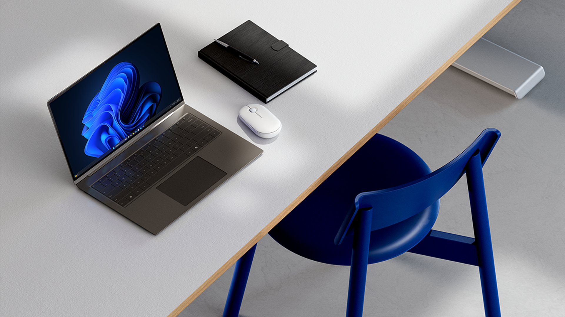 An open laptop displaying a Windows bloom background sits alongside a mouse and notebook on a white desk with a blue chair