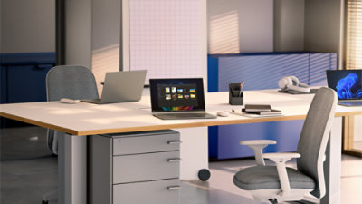 Three open laptops sit on a white desk, alongside notebooks and accessories. One laptop displays a Windows blue bloom background, and one laptop displays open files on top of a Windows rainbow bloom background