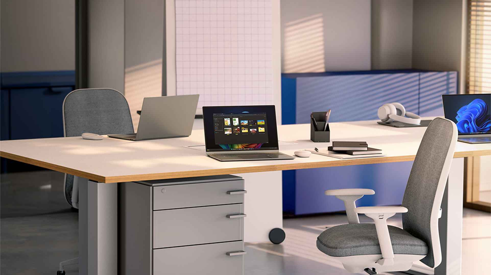 Three open laptops sit on a white desk, alongside notebooks and accessories. One laptop displays a Windows blue bloom background, and one laptop displays open files on top of a Windows rainbow bloom background
