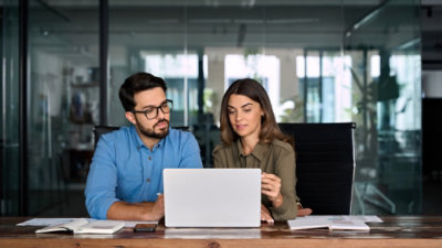 A man and a woman sitting at an office table with a glass wall behind them as they look at a laptop with paper notebooks open and ready on either side