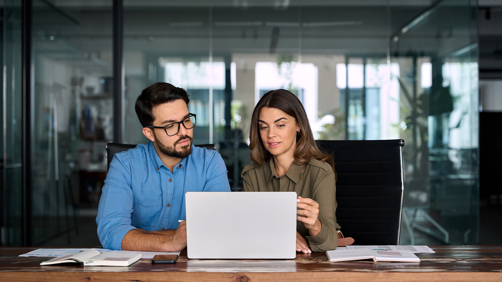 Two office workers sitting at a wood desk in a modern workspace looking at an open Copilot+ PC discussing something onscreen, with notebooks and papers on the desk